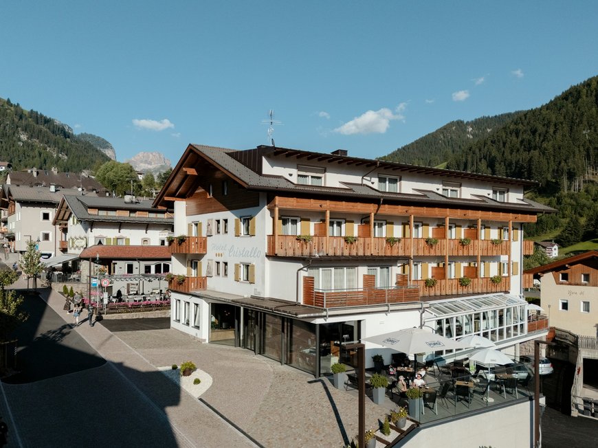 Deine Unterkunft in St. Christina im Grödnertal: Willkommen! Hotelgebäude mit Terrasse und Bergen im Hintergrund unter blauem Himmel