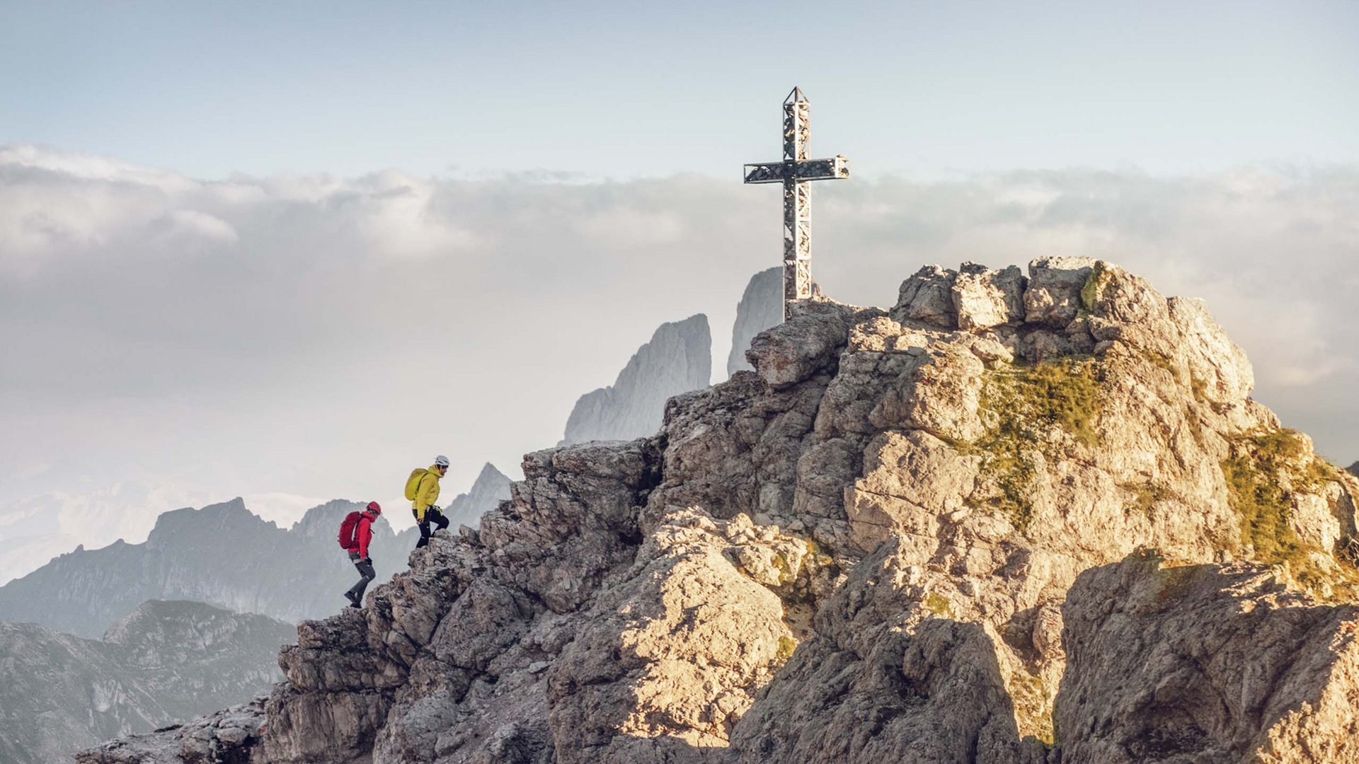 Hotel a Santa Cristina in Val Gardena: your mountain escape Due escursionisti salgono su una montagna rocciosa con croce in vetta
