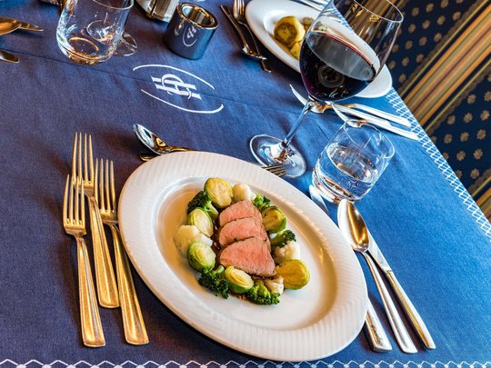 Deine Unterkunft in St. Christina im Grödnertal: Willkommen! Teller mit Fleisch und Gemüse, daneben Glas Rotwein und Wasserglas auf blauem Tischtuch